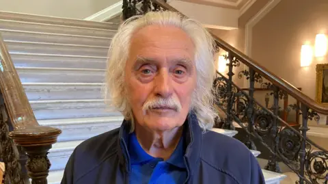 Gemma Dawson/BBC A man with long grey hair and a moustache. He is wearing a blue polo shirt and jacket and is standing at the foot of a large staircase.