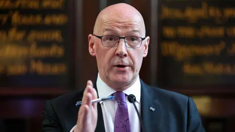John Swinney, who is wearing a navy blazer, a purple tie and a white shirt, addressing a press conference. Behind him, blurred, is a board with calligraphy writing on it.