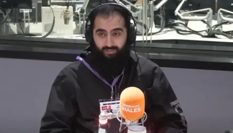 A bearded man wearing headphones sits in a radio studio, speaking into a microphone with an orange “BBC Radio Wales” cover. He is wearing a dark jacket and a lanyard with an ID badge. Studio equipment and microphones are visible behind him through a glass partition.