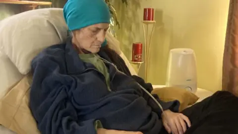 Kelly lies on a hospital bed propped up by pillows and looking down, as if asleep. She wears a blue headscarf on and has a clear tube feeding into her nose. Her hands rest of her lap. 