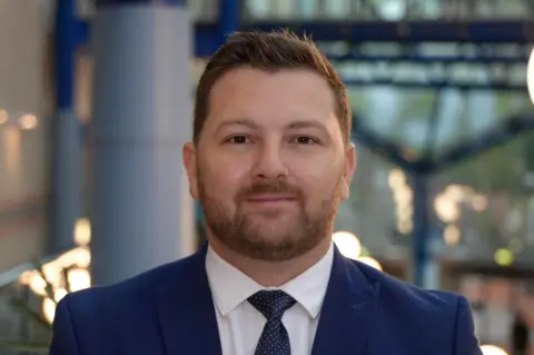 Man in blue suit, with brunette hair, looking directly at the camera.