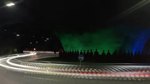 Photograph of vehicles streaking past a roundabout in a tunnel. The tunnel has a green/ blue backdrop and black silhouettes of figures on it.