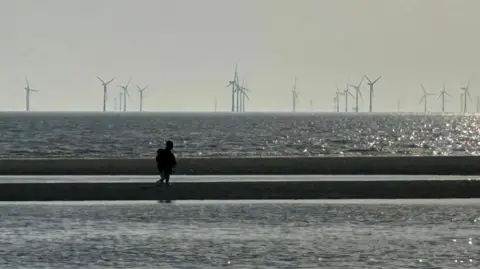 HM Coastguard Crosby A person in silhouette walks along a sand bank on Crosby beach with water pooling on either side and the sea, with an off shore wind farm, in the distance