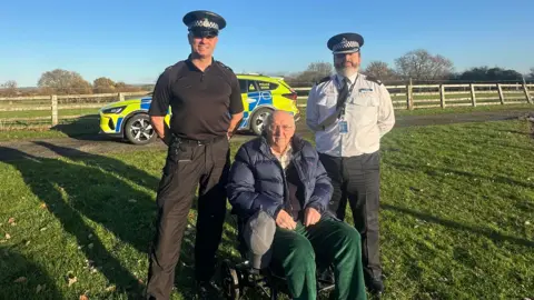 North Yorkshire Police Alan Smith visiting the North Yorkshire Police Dog Section alongside Sgt Gareth Gummerson (left) and Supt Mick Roffe. In the background, a police car can be seen, against a fence with grassy fields behind. Sgt Gummerson and Supt Roffe wear police uniforms and hats. Mr Smith uses a wheelchair and wears a navy blue puffer coat, a black jumper and green cord trousers.