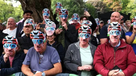 Getty Images Spectators at a cycling event standing behind a barrier and wearing cycling masks which include a man's face wearing blue cycling glasses and a red, white and blue helmet. Everyone is smiling and holding up their phones at the back.