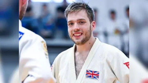 Rowan Kinsella smiling in his white judogi - judo uniform - with a union jack and adaptive judo international badge on it.