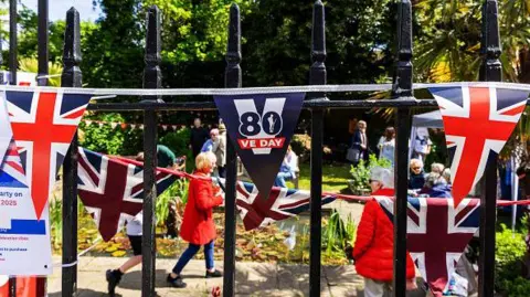Getty Images Flags and bunting are put up on railings for the 80th anniversary of VE Day. People have gathered in a park for a party on a sunny day.