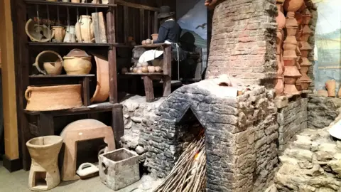 North Devon Council A room full of old pots and a large fire place in a kiln. There is wood stacked inside it and a fire taking place. 