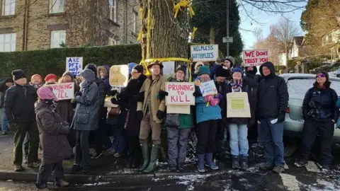 PA Media A small group of around 20 or so protesters gathered around a tree. They are all wearing coats and some of them, hats, on what is a visibly icy day. Some of them hold up placards with slogans such as, "Pruning to kill" and "We don't trust you".