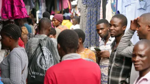 A crowd of people in a market in Lilongwe where clothes stalls can be seen. One person who holds a phone is looking at the camera.