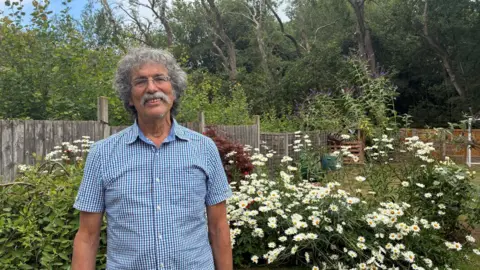 A man with curly grey hair and a grey moustache, wearing glasses and a checked blue and white shirt - stands in front of large white daisy-lie flowers in a garden.
