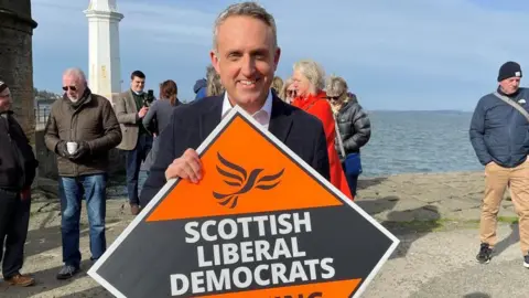 Man on beachfront holding political banner reading "Scottish Liberal Democrats"