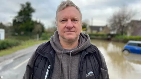 Stuart Portlock, with grey hair and dark clothes,  standing facing the camera with a flooded road in the background