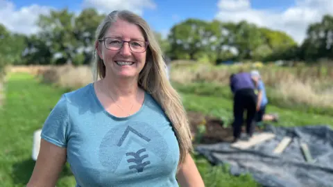 BBC Sheena smiles at the camera as people dig in the field behind her