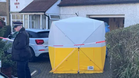 A white and yellow forensics tent outside house and a police officer in uniform