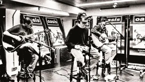 Brian Cannon/Microdot Black and white photo of Bonehead (left), Liam Gallagher (centre) and Noel Gallagher (right) sitting on high stools with microphones in front of them. Bonehead and Noel are playing guitar and Liam is holding a tambourine while singing into the microphone. Large 