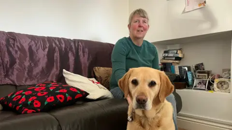 Jenny is sitting on a dark leather sofa with a purple throw and decorative cushions, including one with a red poppy design. In the foreground, a golden Labrador, Nan, sits on the floor. Behind the sofa, there is a recessed shelf filled with books, framed photos, and small decorative items.