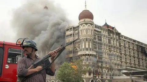 Getty Images Seorang pejabat keamanan India yang mengenakan kemeja bergaris merah dan helm memegang pistol sambil berdiri waspada saat asap dan api mengepul dari bagian hotel The Taj Mahal di Mumbai pada 29 November 2008