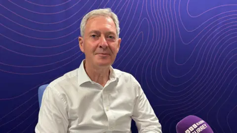Tim Oliver in the BBC Radio Surrey studio. He is wearing a white shirt and looking directly at the camera. The background is dark purple, with swirly light purple lines. The edge of a microphone saying 'BBC Radio Surrey' can be seen on the right hand side.
