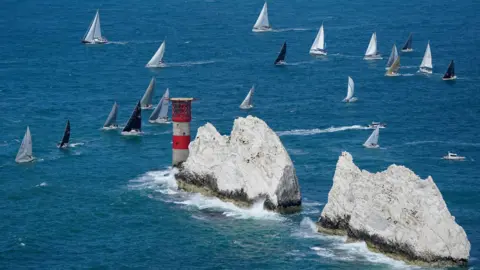 PA Media Needles off the Isle of Wight - large white rocks protruding from the see with a red and white lighthouse. Numerous sailing boats are passing
