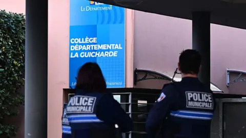 Miguel MEDINA / AFP via Getty Images Two police officers stand in the foreground in front of the school building. A school sign can be seen reading: College Departemental La Guicharde