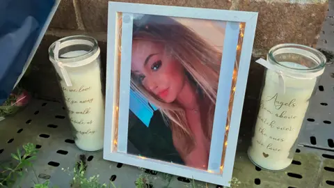 A photo of a young woman inside a white photo frame which has small lights in it. It is propped up against a wall and flanked by two identical white candles.