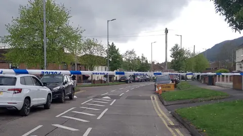 The street has been closed off by blue and white police tape. There are cars parked alongside the street with white road markings up the middle, there is green grass along the side and street lights.