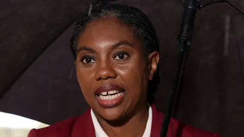 PA Media Headshot of Kemi Badenoch, who has her black hair tied back. She is holding a black umbrella and wearing a burgundy jacket with matching lipstick and a whote collared shirt, without jewellery.