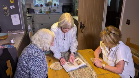 Three women pore over a photo album in a brightly lit kitchen. Two of the women are sat on either side of the table, while the other is stood up and has the album in her hands.