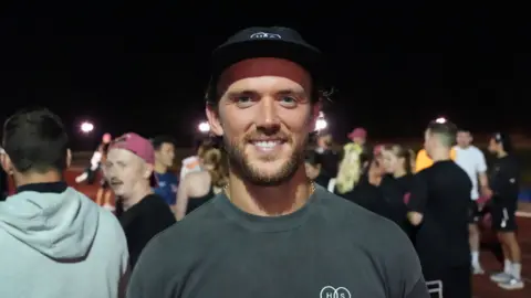 John Fairhall/BBC Emyr Huws is smiling into the camera. He is wearing a grey T-shirt and black baseball cap. Behind him is a crowd of other runners.