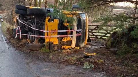 Devon County Council A gritter lorry on its side on a roadside. It has red and white tape around it.