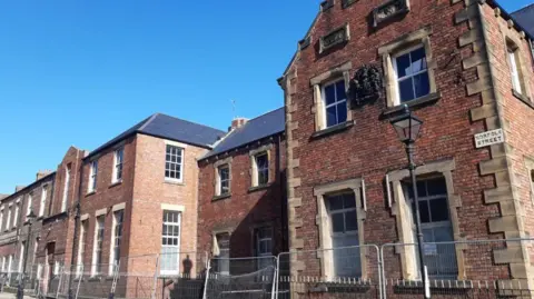 An imposing two-storey red brick building with the Norfolk Street sign. There is metal railing in front of it. 