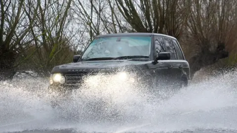 A large 4X4 car drives along a flooded road. Surface water is being catapulted into the air as the car drives through it. The Range Rover is a dark colour and has its headlights turned out. Trees are in the background.