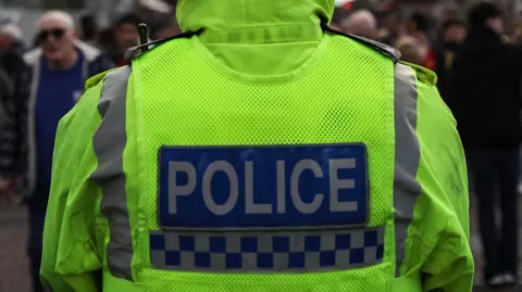 A generic shot of a police officer taken from behind. He is wearing a hi-viz jacket with the word 'police' printed on the back.