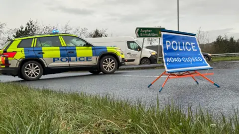 A police car is parked in the middle of a road, restricting entry. There is a blue rood sign which reads "Police road closed". There is a green road sign in the background which is pointing to the towns of Augher and Enniskillen. 