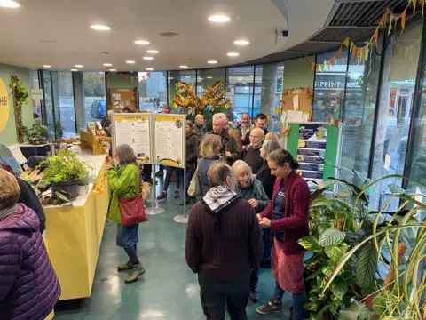 Climate Hub BANES Circular room with windows looking out onto a street in Bath. People are in discussion in pockets around the room, and there are yellow and white displays boards that tell people about a particular climate issue.