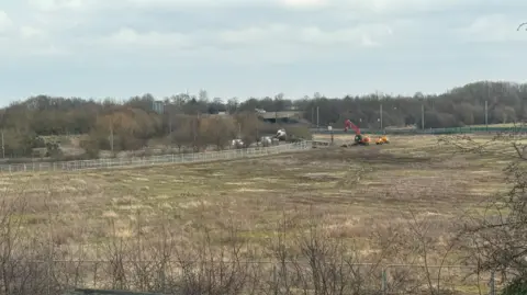 SUPPLIED A large field with a metal fence and digging machine in the middle distance ahead of a tree line and road bridge.