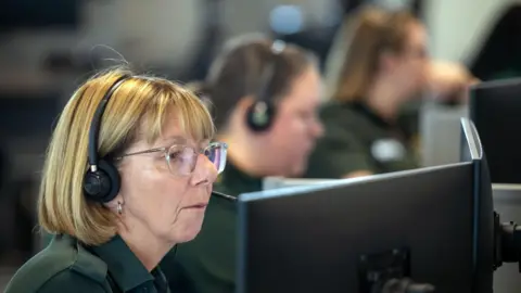 A blonde woman wearing glasses and a green polo shirt. She is sitting inside an ambulance call room with several others.