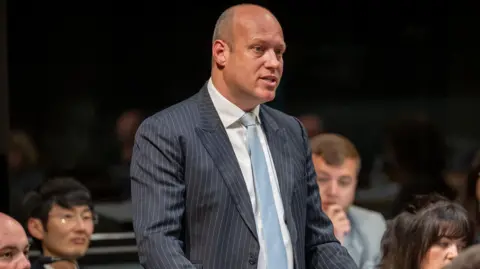 Senedd Cymru Dan Thomas speaking in a pinstriped suit with a white shirt and blue tie/