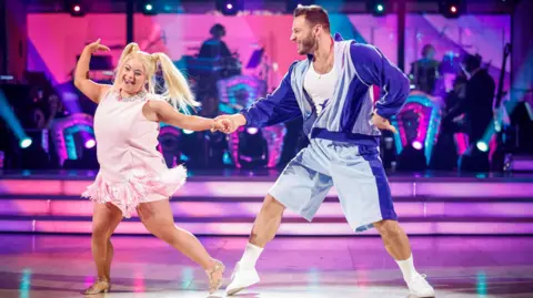 Ellie and Vito dancing in the Strictly studio with the band behind them. Ellie is dressed as Emma Bunton, wearing a pink dress and a blonde wig with bunches. Vito is dressed in a white vest, blue tracksuit jacket and matching shorts
