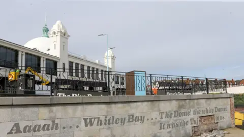 The area outside the Spanish City Dome in Whitley Bay. The area is a viewing point with black railings and an inscription along the front wall of the platform. Behind it there is a large white pavilion building with a dome and two turrets.