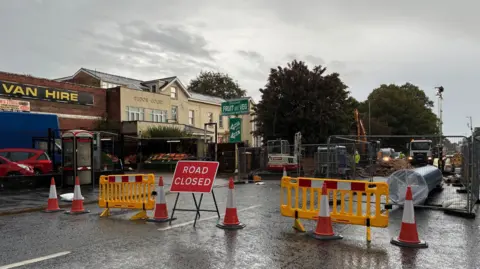 Road Closed sign next to traffic cones and yellow fencing - with fencing around a hole where repairs take place