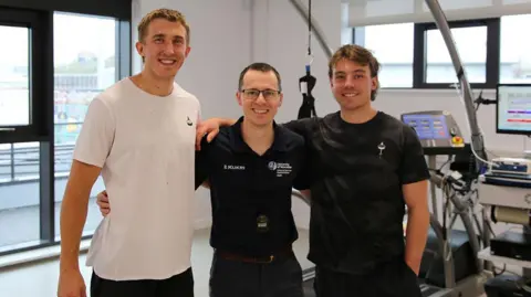 Alfie (Left) and Joe Pritchard (Right) with Dr Matt Cook (Centre). The man on the left is wearing a whit t-shirt and black trousers. The other two men are wearing black t-shirts and dark trousers. All are smiling at the camera and there is gym equipment behind them. 