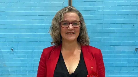 A woman with shoulder length, curly blonde hair, stands in front of a blue-painted brick wall. She wears a red jacket over a black top and has red-rimmed glasses.