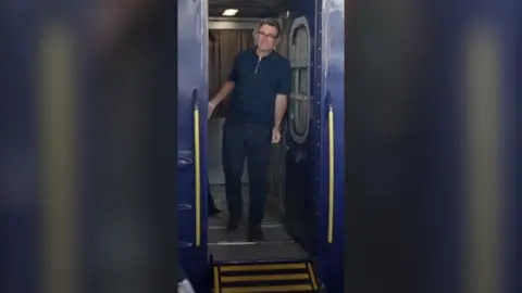 UK Prime Minister Andy Burnham wearing black t-shirt and trousers stepping out of train