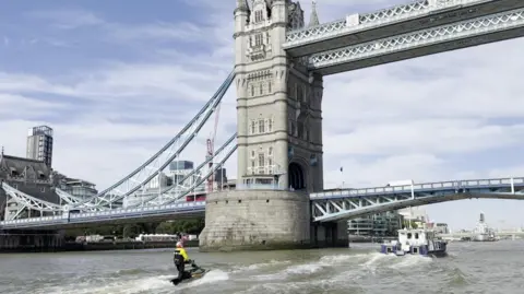 Lee Robinson A jet skier wearing a yellow waterproof jacket rides a black stand-up jet ski across the River Thames in London, passing directly in front of Tower Bridge with an escort boat following behind.