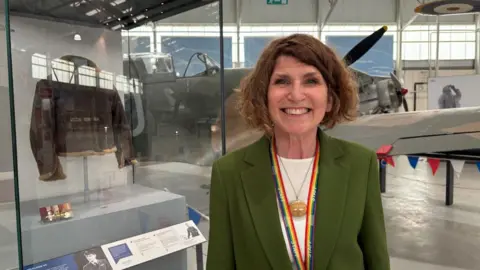 A woman with wavy bobbed brown hair, wearing a a green jacket, white top and necklace, is standing in a large open space with planes on display behind her. She has a rainbow striped lanyard around her neck.