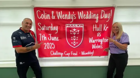 A red sign with white writing reading "Colin & Wendy's Wedding Day! Saturday 7th June 2025. Hull KR vs Warrington Wolves. Challenge Cup Final - Wembley!" with a Hull KR logo in the middle. White blinds are pulled down behind the sign and the wall below is painted dark green. Colin is standing on the left and Wendy is standing on the right. They are both pointing to the flag. Colin is wearing black trousers and a blue and red rugby short-sleeve top. He has short grey hair. Wendy is wearing black trousers with a purple short-sleeve rugby top. She has shoulder-length straight blonde hair. 
