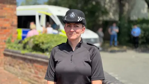Insp Liz Warner pictured wearing a black police top with a quarter zip and a black police hat. Behind her in the blurred background are people in plain clothes stood next to a police van.