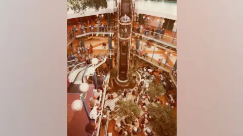 Other A busy multi‑level indoor shopping centre photographed from an elevated viewpoint. At the centre is a dome‑shaped lift. Several floors of walkways, railings and escalators surround the lift, with people standing, walking or watching from the balconies.